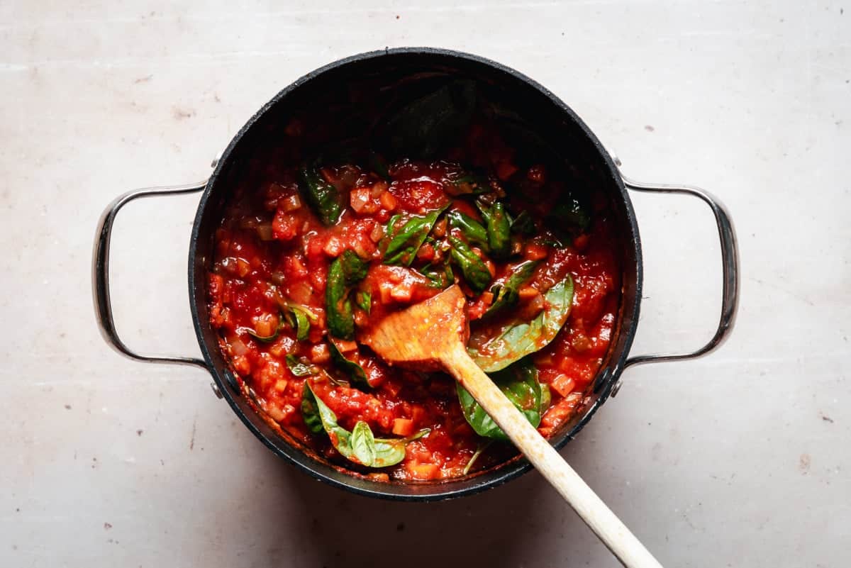 A top view of a black pot containing a vibrant tomato soup mixed with fresh basil leaves. A wooden spoon rests inside the pot, surrounded by a light-colored countertop.