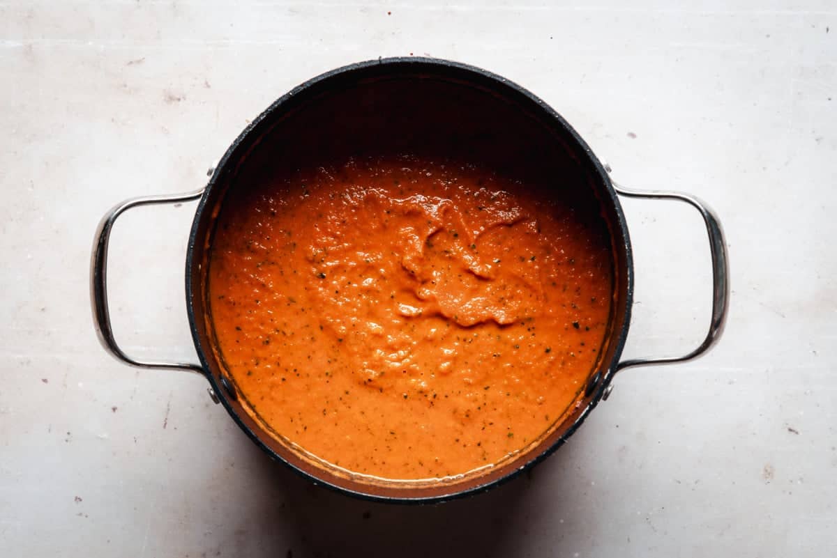 A pot filled with thick, orange-red tomato soup with visible herbs on a light-colored surface. The soup is slightly textured and appears freshly made. The pot has handles on both sides.