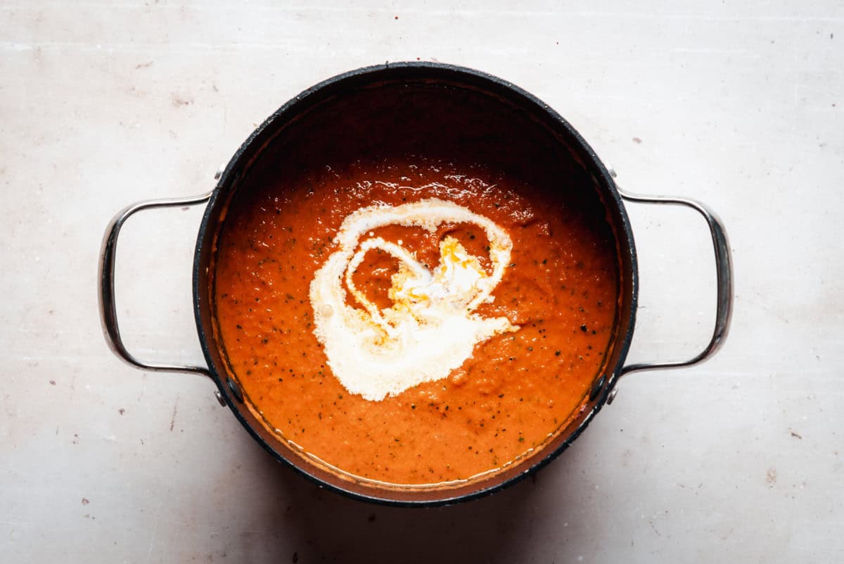 A pot of tomato soup with a swirl of cream on top sits on a light surface. The soup is vibrant orange-red, and the cream adds a contrasting white design in the center. Silver handles extend from both sides of the pot.