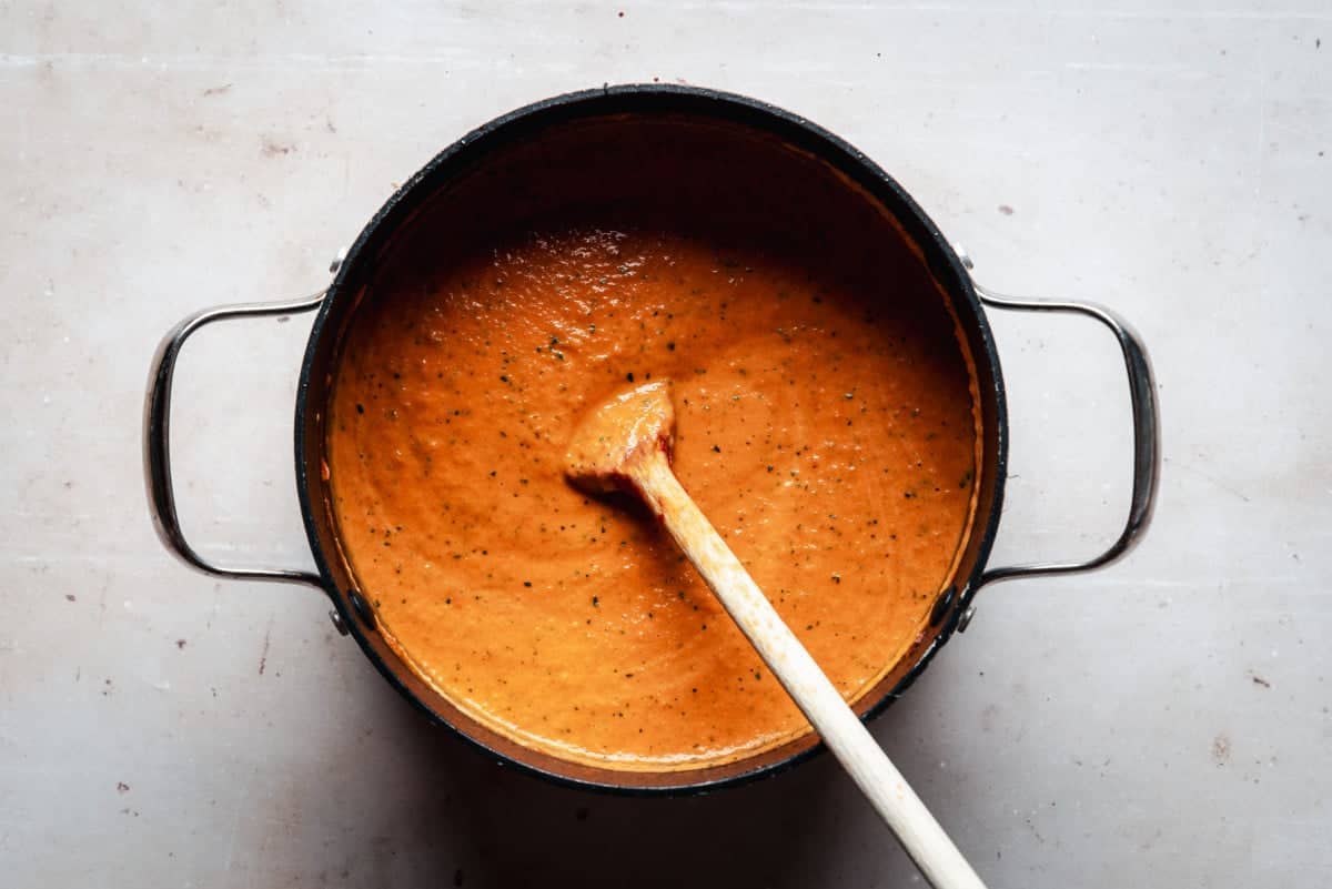 A black pot filled with creamy tomato soup sits on a light surface. A wooden spoon rests inside the pot, slightly tilting to the side. The soup appears smooth with small herb flecks visible.