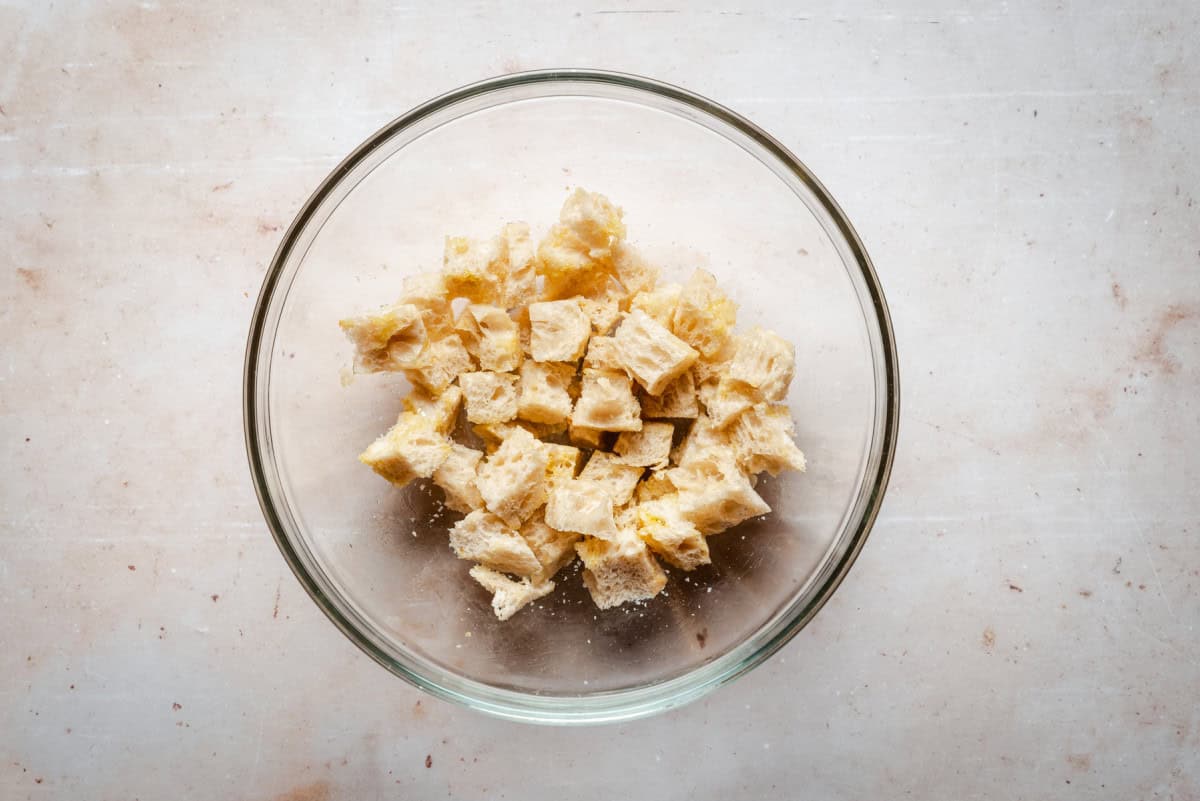 A glass bowl filled with small, cubed pieces of bread, lightly coated in olive oil and salt on a textured, light-colored surface.