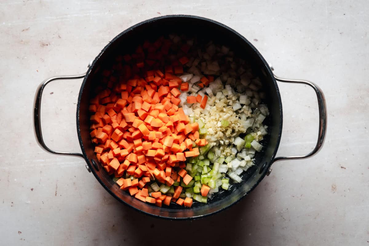 A pot filled with chopped carrots, celery, onions, and garlic sits on a light countertop. The colorful vegetables are neatly arranged, ready to be cooked.