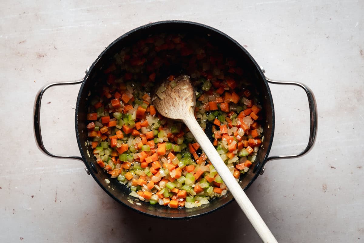 A black pot filled with diced carrots, celery, and onions, being stirred with a wooden spoon. The pot is on a light-colored surface.