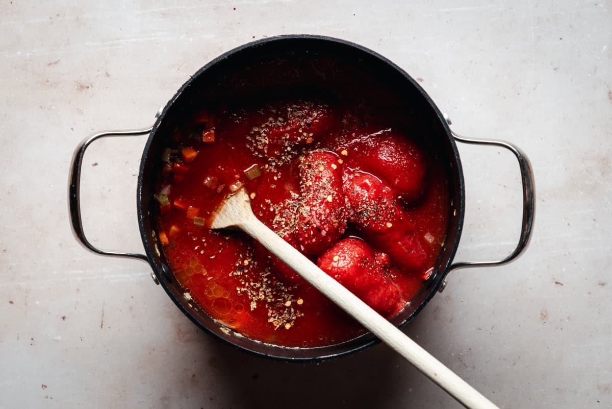 A pot of simmering vegetables with whole tomatoes and spices added, being stirred with a wooden spoon, on a light-colored surface.