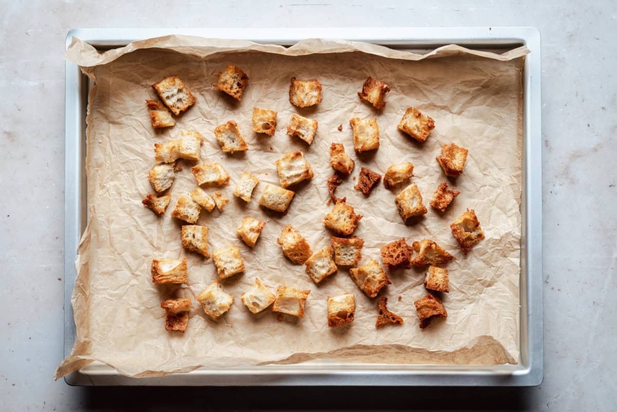 A baking tray lined with parchment paper holds evenly spaced, golden-brown croutons. The croutons are arranged randomly, showcasing their crispy texture.