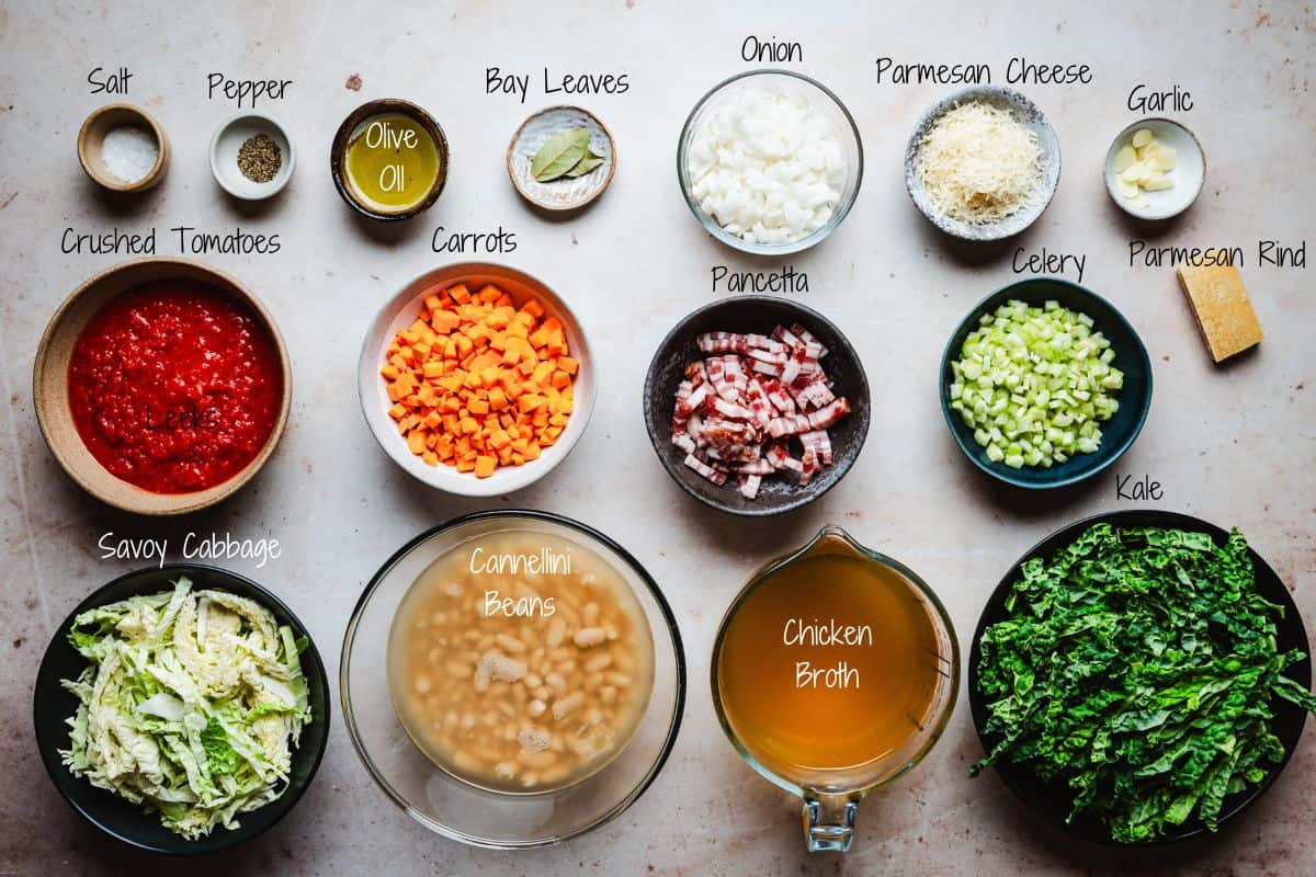 Ingredients are laid out for making Tuscan White Bean Soup, including seasonings, olive oil, bay leaves, chopped onion, parmesan cheese, garlic, crushed tomatoes, carrots, pancetta, celery, parmesan rind, savoy cabbage, cannellini beans, chicken broth, and kale.