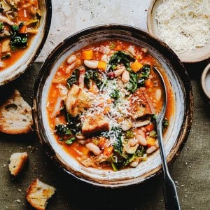 A bowl of hearty vegetable soup with kale, white beans, and chunks of bread topped with grated cheese. A spoon rests inside the bowl. There are pieces of crusty bread and a small bowl of cheese nearby on a rustic surface.