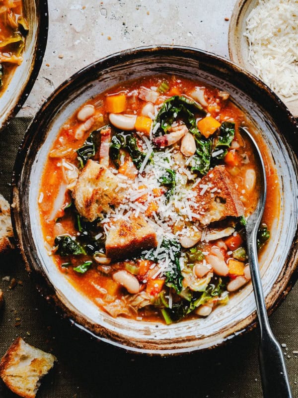 A bowl of hearty vegetable soup with kale, white beans, and chunks of bread topped with grated cheese. A spoon rests inside the bowl. There are pieces of crusty bread and a small bowl of cheese nearby on a rustic surface.