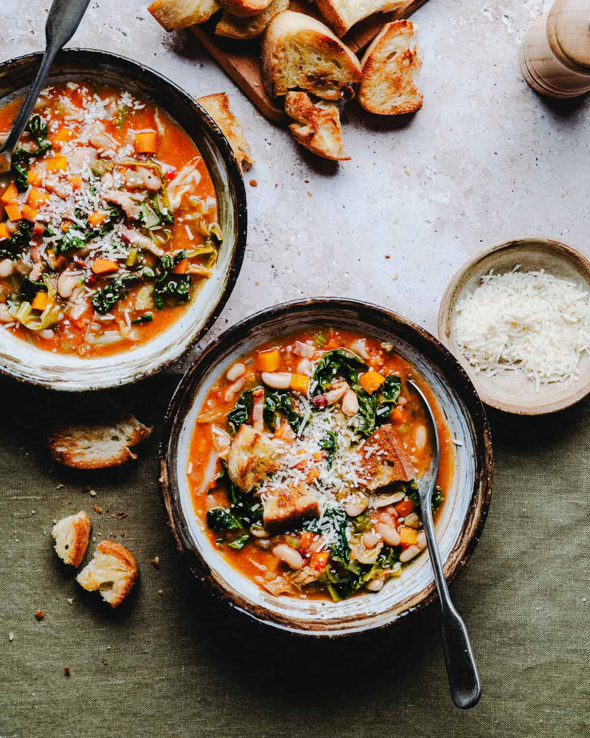 Two bowls of soup topped with grated cheese, placed on a textured surface. Nearby are scattered slices of toasted bread and a small bowl of more grated cheese. The scene conveys a cozy, rustic meal setting.