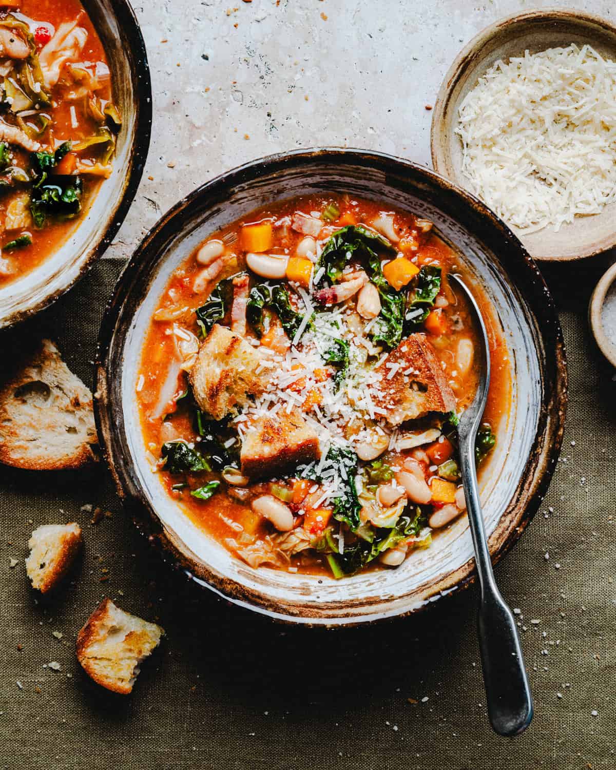 A rustic bowl of hearty vegetable soup featuring beans, kale, and tomatoes, topped with grated cheese and crusty bread. A fork rests on the side of the bowl, with additional bread pieces and a bowl of grated cheese nearby.