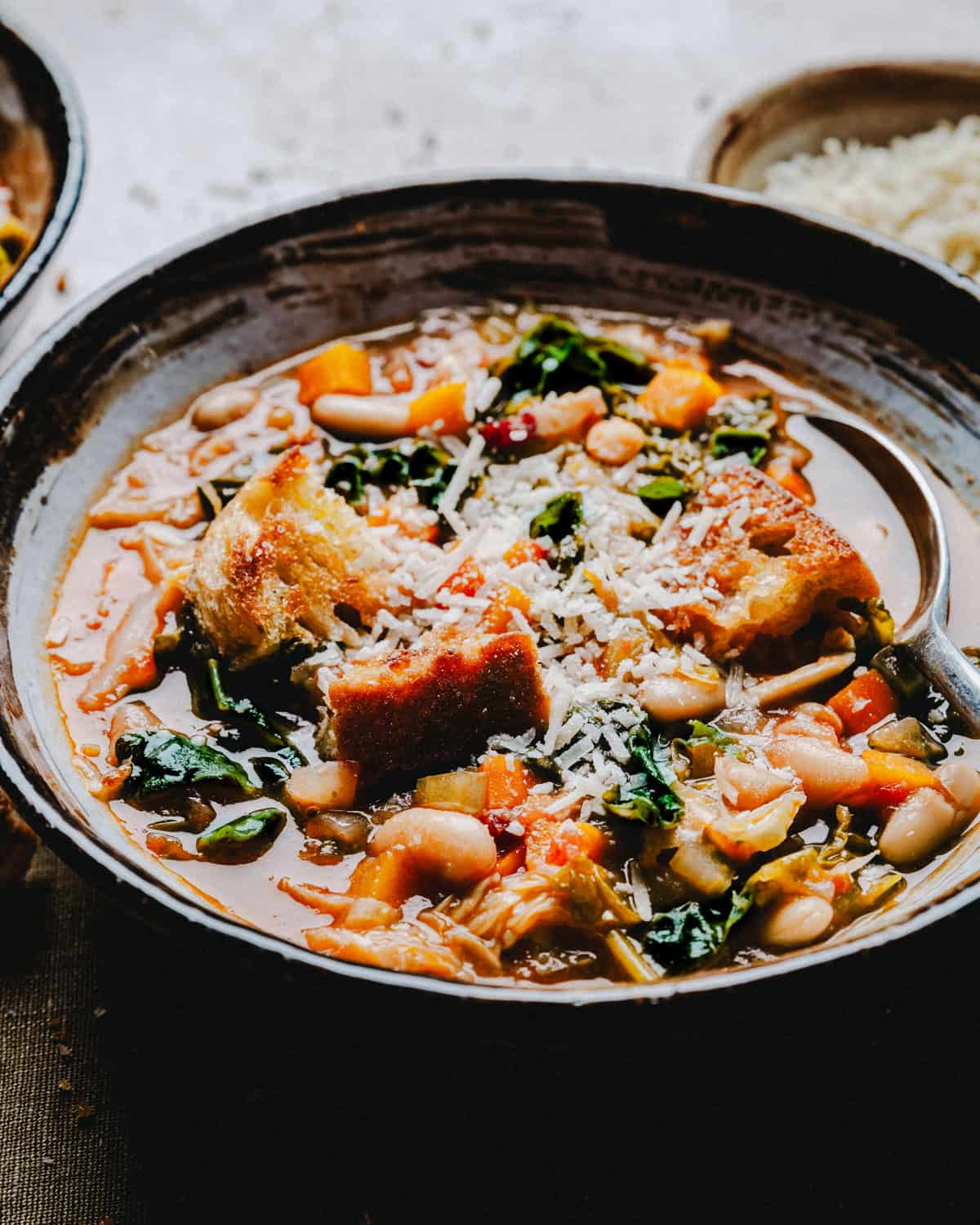 A hearty bowl of vegetable and bean soup topped with crispy, toasted bread pieces and garnished with grated cheese. The soup is filled with colorful vegetables and leafy greens, accompanied by a spoon resting on the side of the rustic bowl.