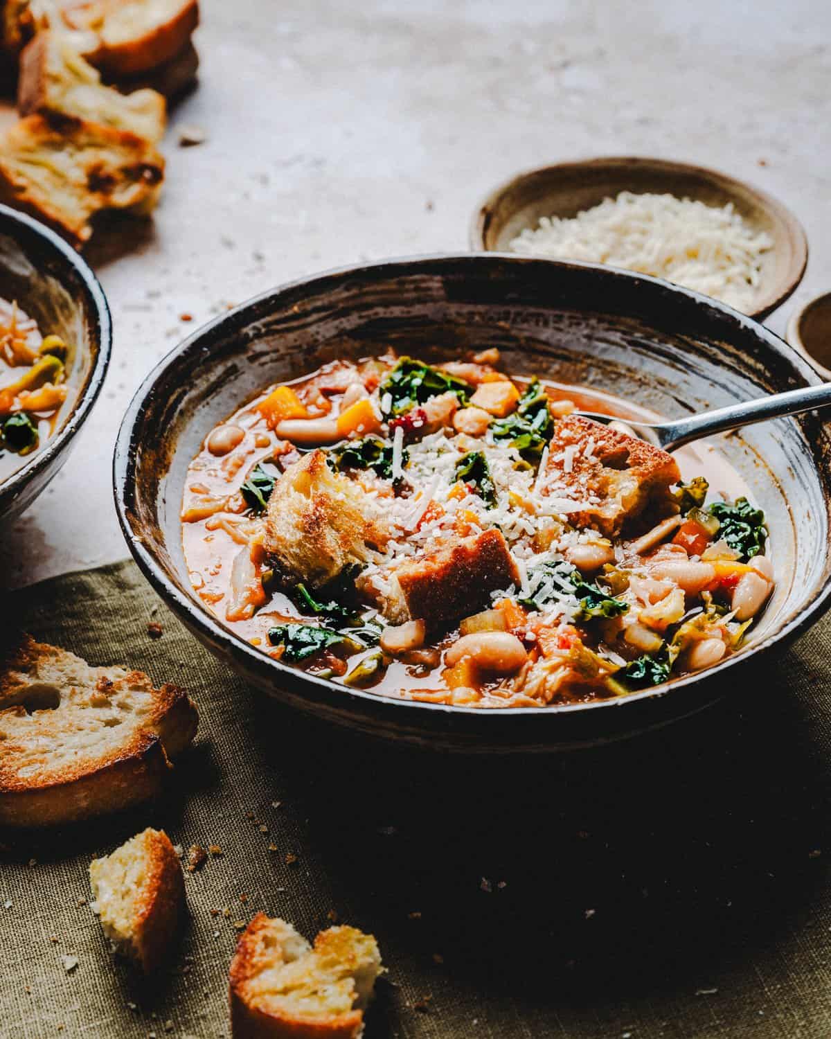 A hearty bowl of vegetable and bean soup garnished with grated cheese and served with slices of crusty bread. The soup includes greens and appears warm and comforting, creating a rustic and inviting dining scene.