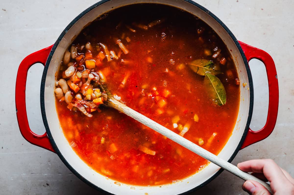 A red pot filled with broth filled soup, including carrots, beans, and leafy greens, being stirred with a wooden spoon. The broth is a rich tomato color, and the pot rests on a light-colored surface.