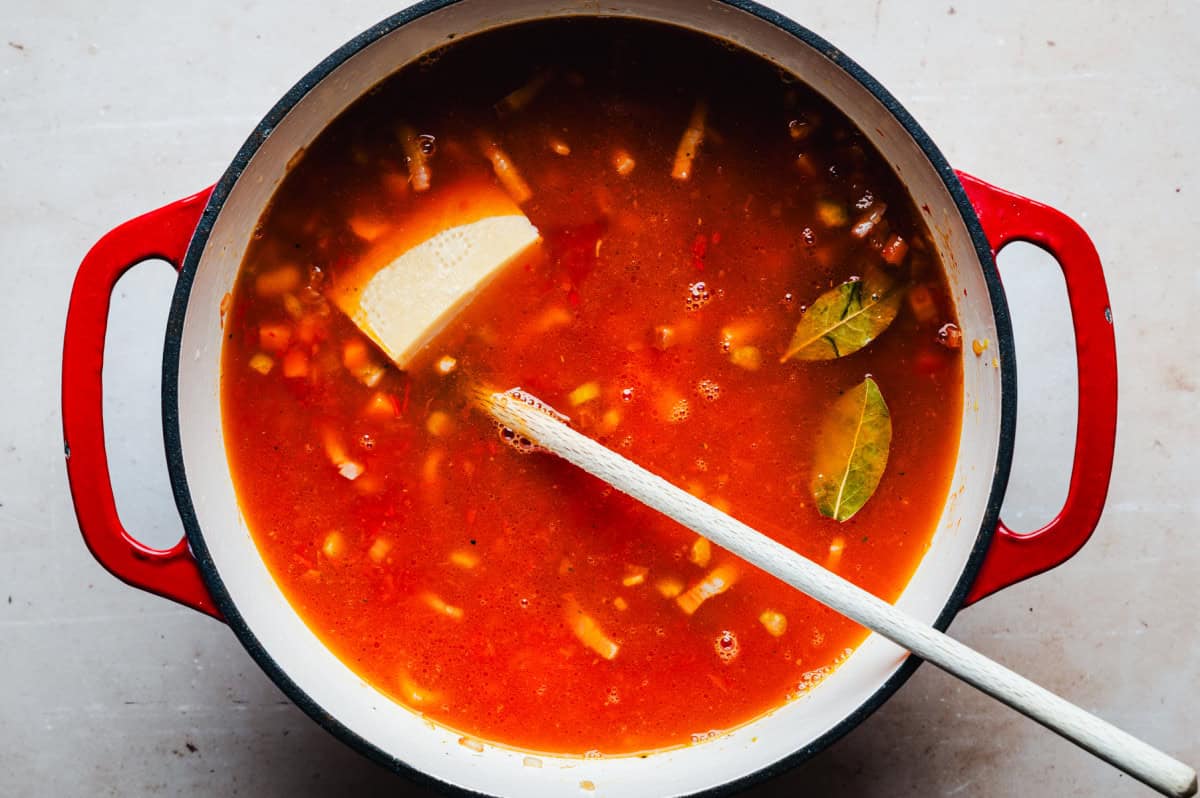 A pot of tomato-based soup with a wooden spoon inside, containing cubes of parmesan rind, bay leaves, and chopped vegetables. The pot has red handles and rests on a light-colored surface.