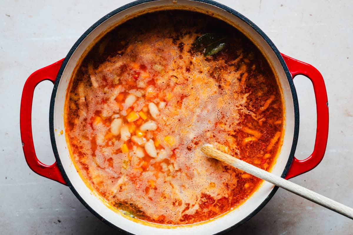 A red pot filled with a bubbling vegetable soup, featuring chunks of carrots, tomatoes, and beans. A wooden spoon rests inside, stirring the vibrant, simmering broth. The pot sits on a light surface.