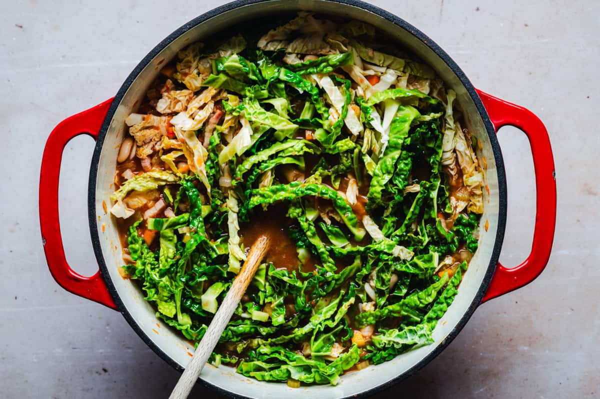 Top view of a red pot filled with a simmering soup. The ingredients include chopped leafy greens, sliced cabbage, and a rich broth. A wooden spoon rests in the pot, suggesting the dish is being stirred or served.