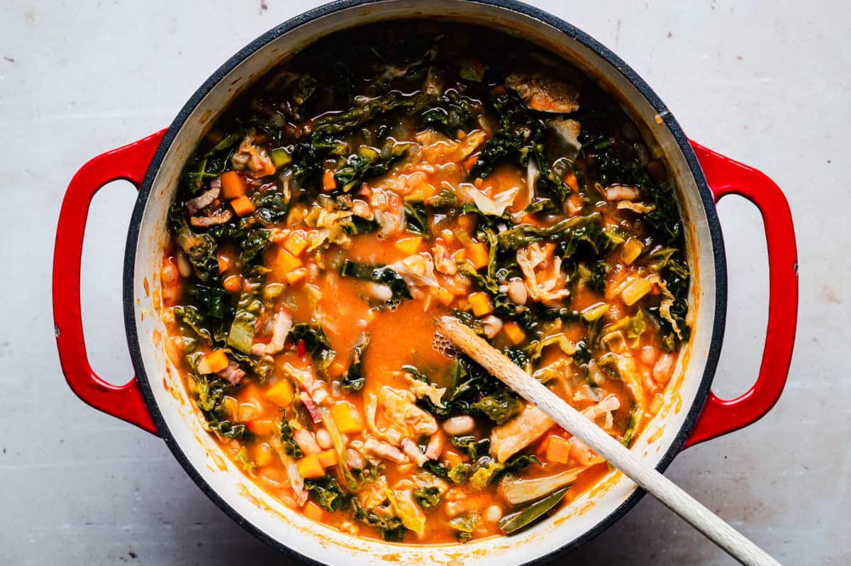 A red pot filled with a hearty soup containing sliced carrots, kale, white beans, and bits of pancetta in a rich tomato broth. A wooden spoon rests inside the pot, suggesting it's ready to serve.