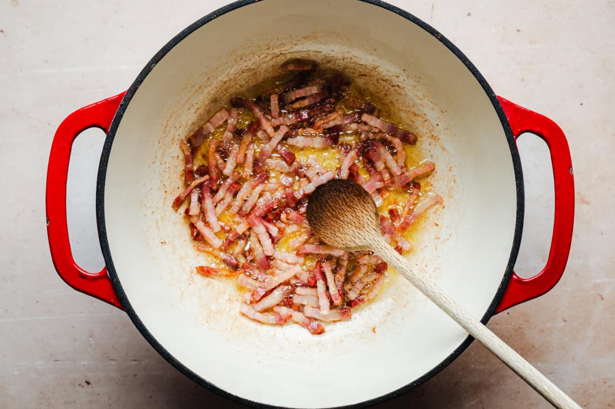 A red-handled pot on a light surface contains sizzling strips of pancetta being cooked with a wooden spoon. The bacon is browning, releasing fat and flavors.