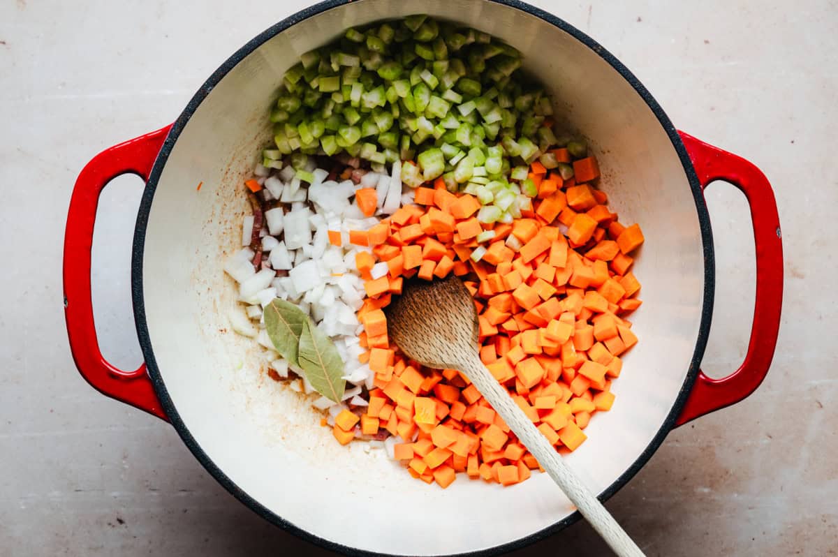 A pot with a wooden spoon contains chopped carrots, celery, onions, and bay leaves. The pot has red handles and is placed on a light surface.