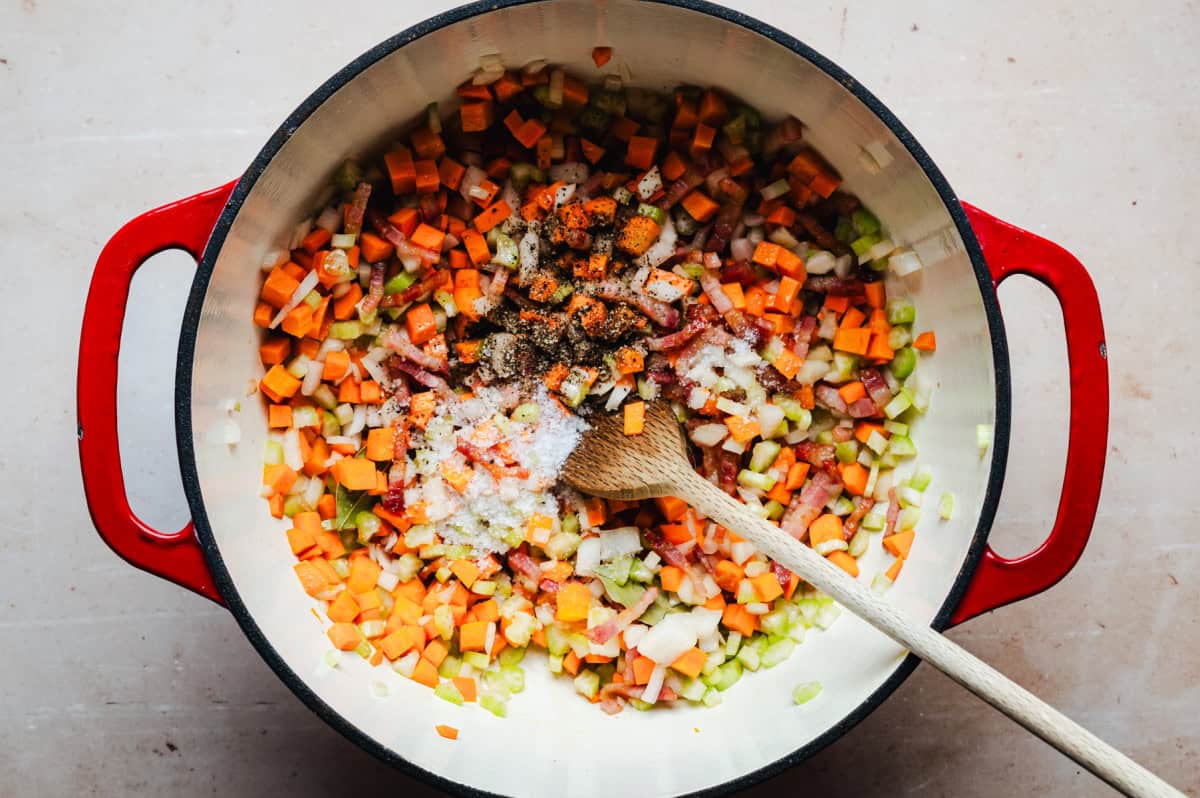 A red pot filled with chopped vegetables including carrots, onions, and celery, being stirred with a wooden spoon. Black pepper are sprinkled on top. The mixture is placed on a light-colored countertop.