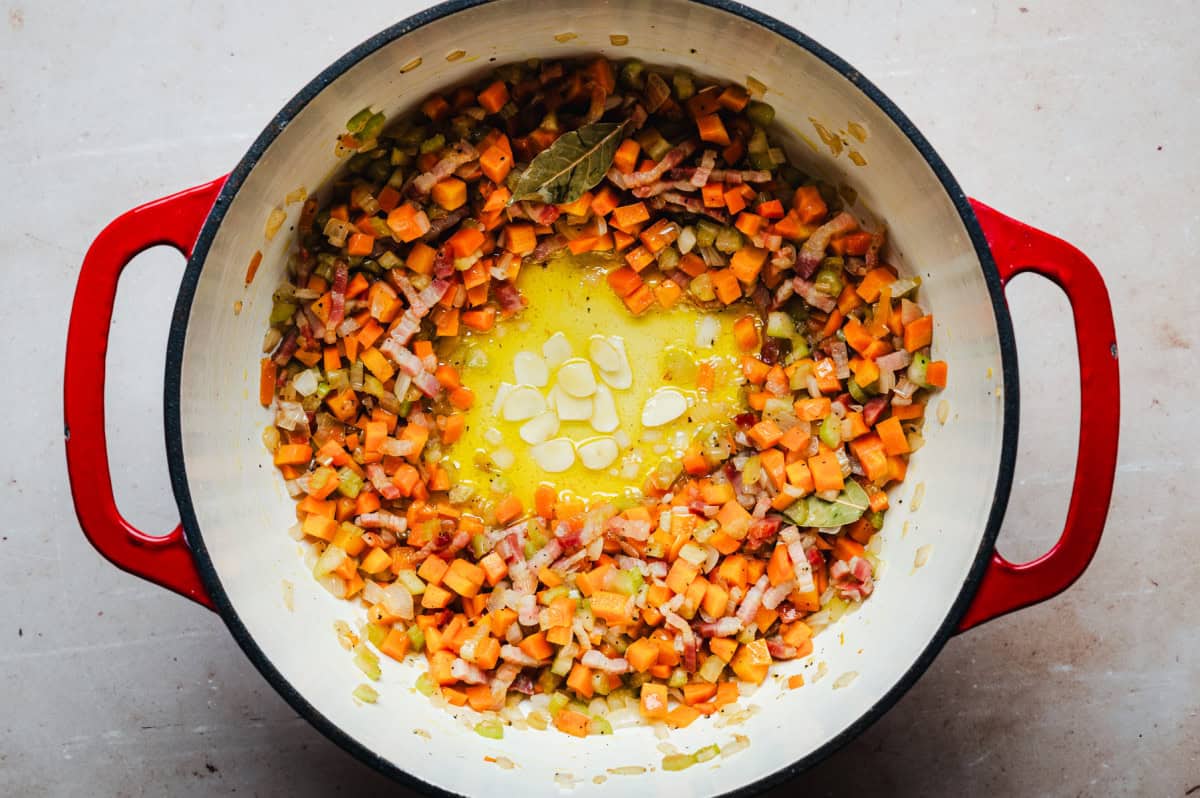 A red pot on a stovetop contains chopped carrots, celery, and bacon sautéing in melted butter with sliced garlic and bay leaves for seasoning.