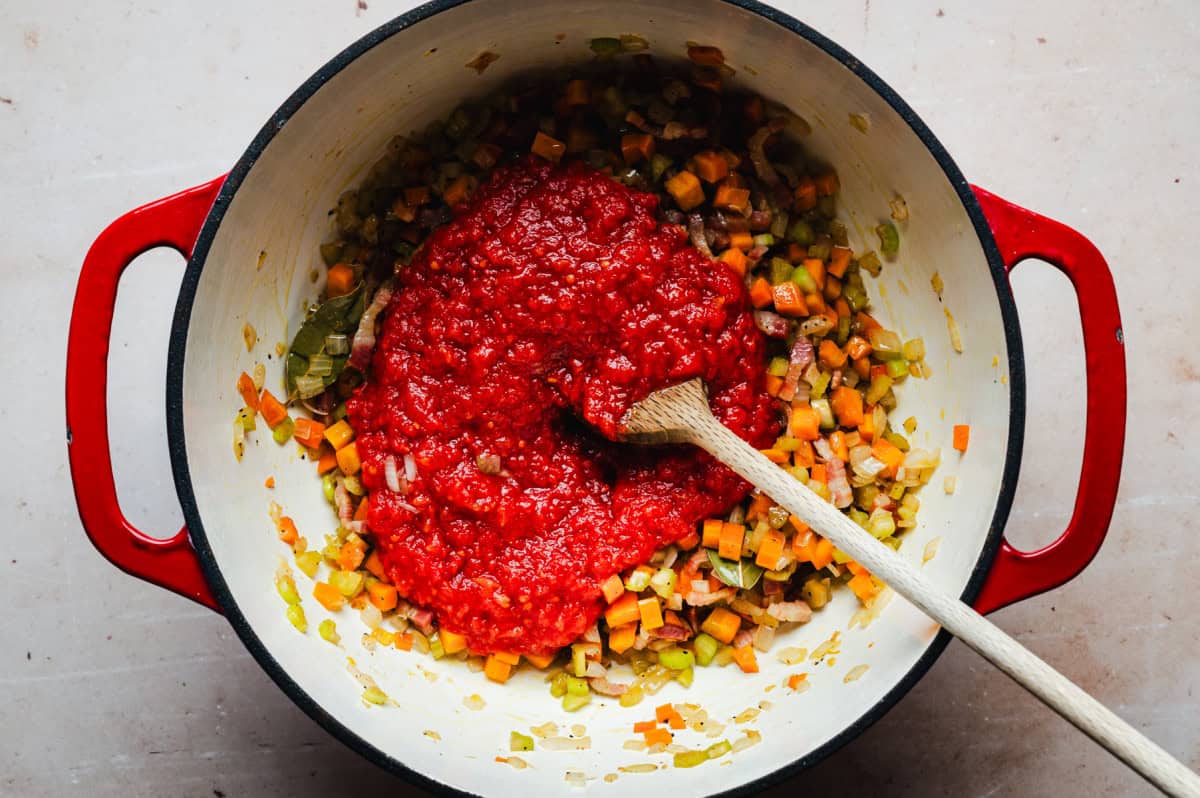 A red pot filled with chopped vegetables, including carrots, celery, and onions, sautéed and topped with crushed tomatoes. A wooden spoon rests inside, stirring the ingredients. The pot is on a light-colored surface.