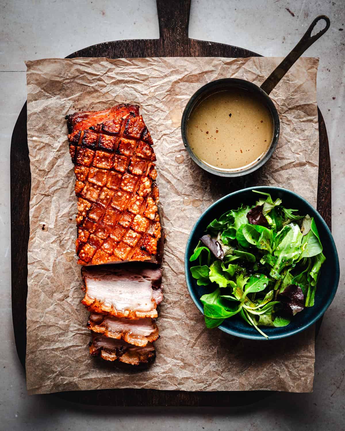 Slab of cooked, crispy skin on pork belly on a cutting board. A few slices cut from and next to a simple green salad.