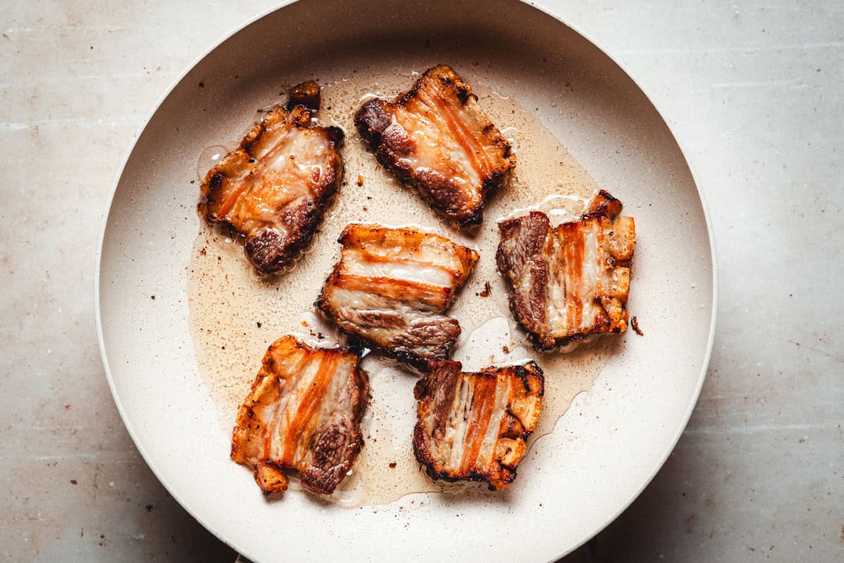 Slices of pork belly frying in oil in a stovetop skillet.