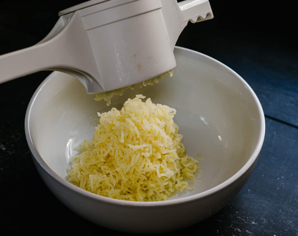 pressing steamed yukon gold potatoes through potato a ricer into a white bowl.